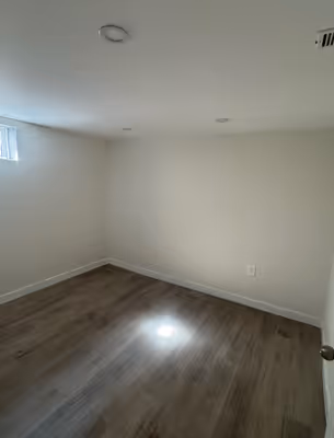 A similar view to the first image, showing an empty room with white walls, light wooden floors, and a doorway.