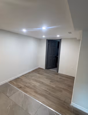 A wider view of the empty room, showcasing the light wooden floors, white walls, and a doorway leading to another room with similar finishes.