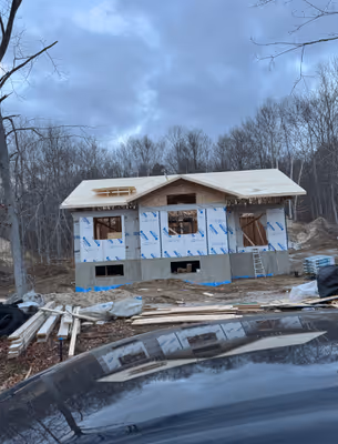 An exterior shot of a partially constructed house with visible wooden framing, set against a backdrop of trees and a cloudy sky.