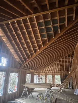 Wide interior shot of a building under construction, showing the exposed wooden beams of the roof structure and the open space below.